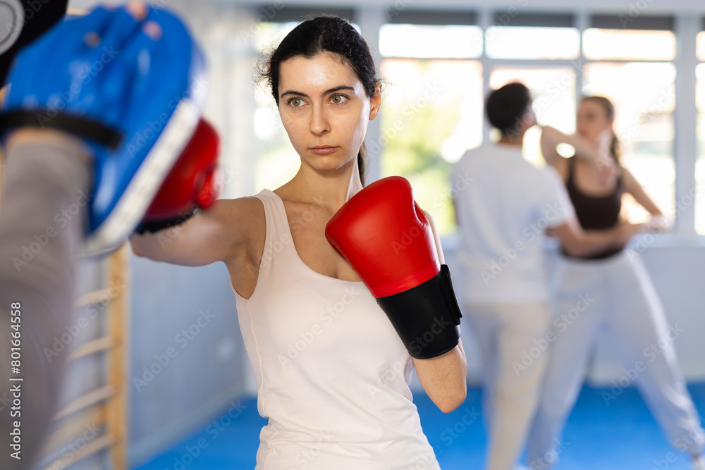 Sportive young female practitioner of boxing courses applying kicks on hitting mitts during workout session