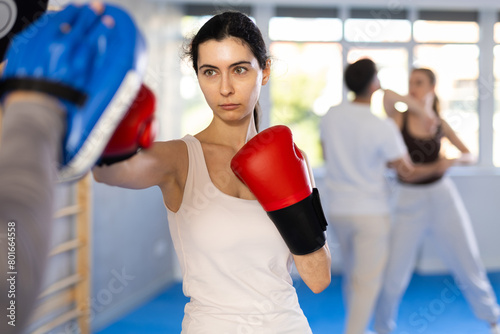 Sportive young female practitioner of boxing courses applying kicks on hitting mitts during workout session © JackF