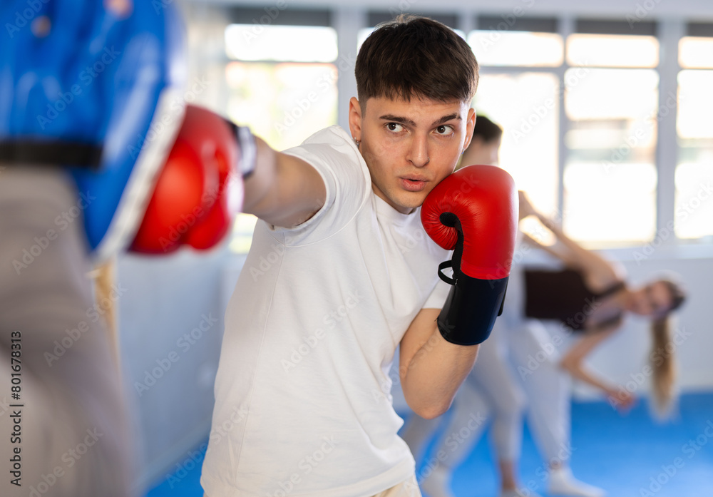 Sportive young male practitioner of boxing courses applying kicks on punch mitts during workout session