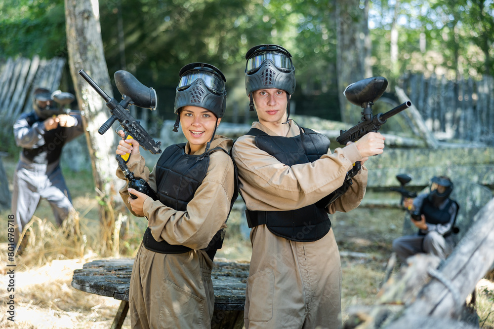 Fototapeta premium Cheerful boy and girl wearing protective paintball equipment and helmets, holding guns and posing.