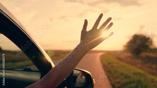 Young woman driver rides car catches wind with her hand from car window. Girl with long hair sits in front seat of car, her hand out window catching wind, glare of setting sun. Child travels by car photo