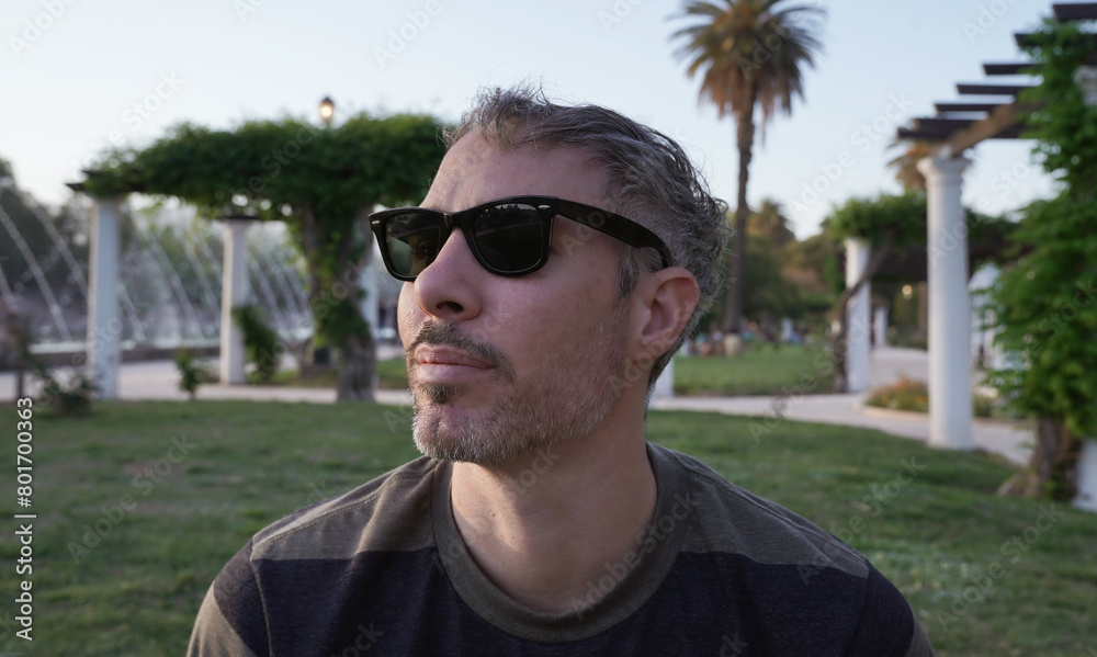 Portrait of a man in his 30s, wearing sunglasses sitting in the grass in the park at sunset. The white pergola columns in the background.
Portrait of a man in his 30s, wearing sunglasses sitting in the grass in the park at sunset. The white pergola columns in the background.