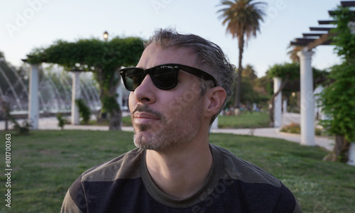 Portrait of a man in his 30s, wearing sunglasses sitting in the grass in the park at sunset. The white pergola columns in the background.	
