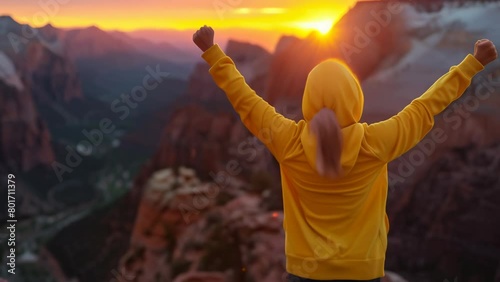 Woman in yellow hoodie stands on mountaintop at sunset, raising arms triumphantly, face and landscape illuminated by warm glow, evoking sense of accomplishment and awe-inspiring beauty.
