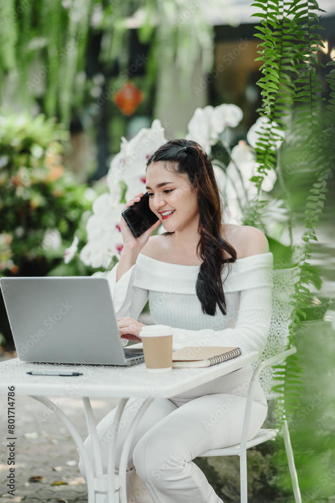 Smiling young woman takes a phone call while working on her laptop at a peaceful garden cafe, multitasking with ease. Smiling young woman takes a phone call while working on her laptop at a peaceful garden cafe, multitasking with ease.