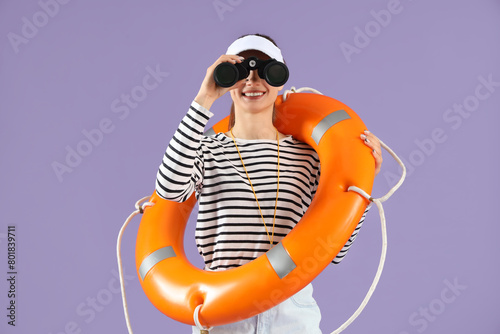 Young female lifeguard in cap with rescue ring looking in binocular on lilac background
