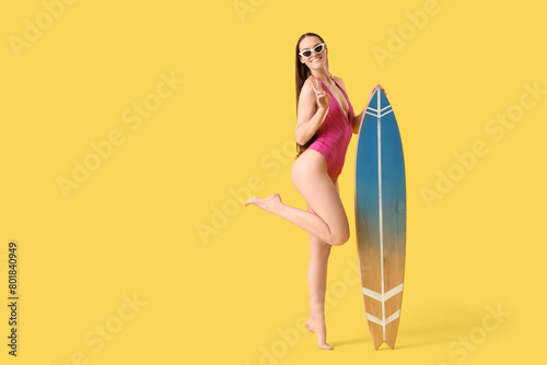 Smiling woman with sunglasses and surfboard showing victory gesture on yellow background
