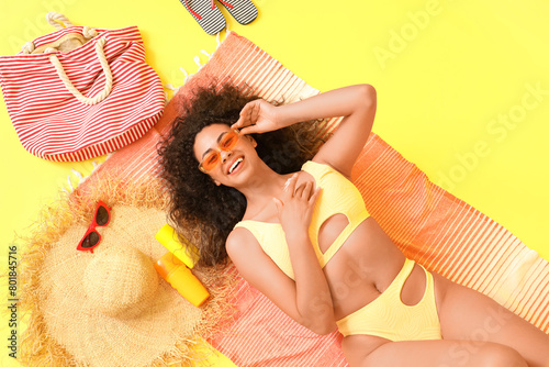 Young African-American woman applying sunscreen cream on beach towel against yellow background, top view