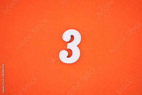 The number 3 is made from white painted wood placed on a background of orange paper.