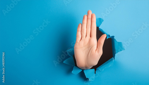 Hand making a stop sign, coming out of a torn blue paper hole. Concept of stop photo