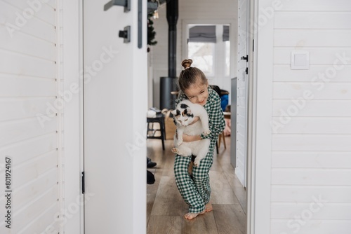 A little blond girl wearing pajamas hugs a domestic cat in her bedroom. The concept of a cozy home morning