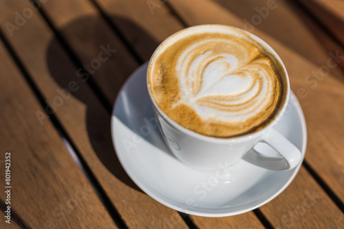 Coffee on wooden background. Cup of cappuccino with latte art on brown table with sunbeam. Fresh morning coffee with delicious milk foam.