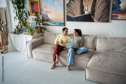 Cheerful couple spouses relaxing on couch after working day. Man with mug, woman with laptop, loving smiling couple having conversation discussing communicating in house studio among art paintings
