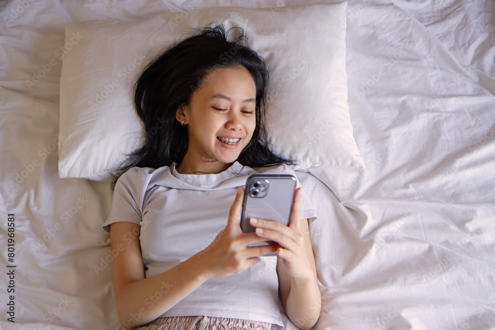 Top view of happy asian young woman awaken in white cozy bed using modern smartphone gadget, happy millennial female wake up lie in comfortable bedroom browsing Internet on cellphone in the morning Top view of happy asian young woman awaken in white cozy bed using modern smartphone gadget, happy millennial female wake up lie in comfortable bedroom browsing Internet on cellphone in the morning