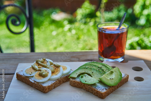 Two open sandwitches from wholegrain bread spread by fresh cheese and decorated by healthy,  balanced ingredients like slices of fresh avocado and boiled eggs and served outside with cup of black tea.