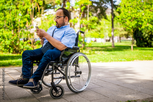 Portrait of man in wheelchair. He is enjoying sunny day in city park and messaging on smartphone.