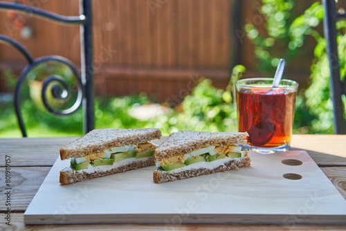 Healthy and blanced breakfast wholegrain sandwich splitted in two, spreaded with cheese and slices of avocado and hard boiled egg served outside in the garden on the wooden plate with cup of black tea
