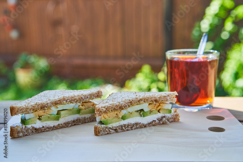 Healthy breakfast served outside in the garden restaurant in the sunny summer morning. Wholegrain sandwich splited in two spreaded with fresh cheese and slices of boiled egg and avocado with black tea