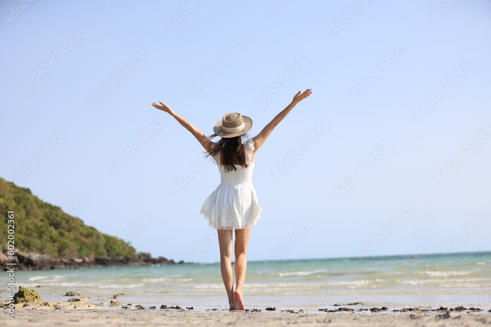 Young attractive ethnic Thai millennial woman posing at suny spreading arms to sides and various postures dressed in white casual clothes stands on the ocean with sandy beach. Love for travel Young attractive ethnic Thai millennial woman posing at suny spreading arms to sides and various postures dressed in white casual clothes stands on the ocean with sandy beach. Love for travel