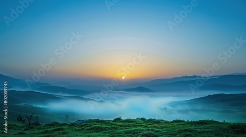 a foggy landscape. The foreground is a green grassy field with some yellow flowers. In the background are hills and trees covered in a thick white fog.  © Awais