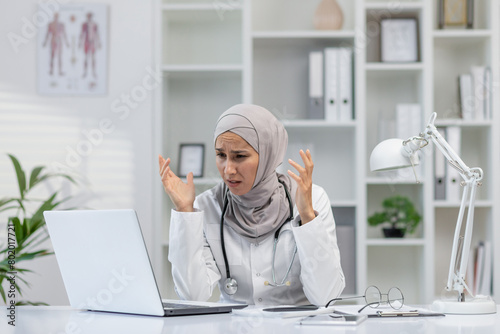 A female doctor wearing a hijab shows signs of frustration and confusion while using her laptop in a bright medical office. Her expression conveys stress and difficulties.