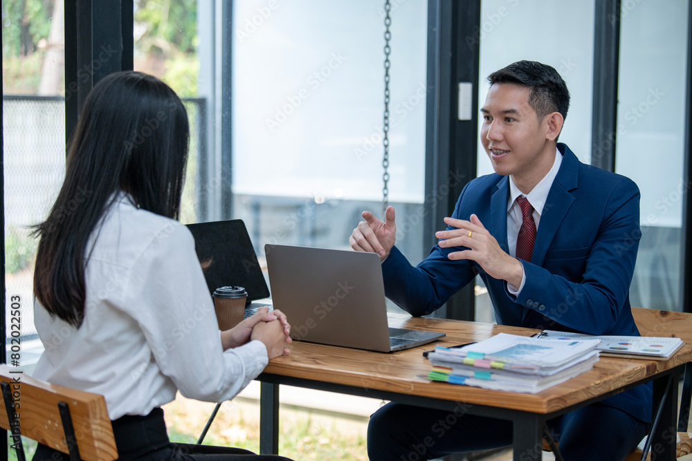 Two Asian professionals engaging in a thoughtful discussion at a business meeting, with a laptop and documents on the table. Two Asian professionals engaging in a thoughtful discussion at a business meeting, with a laptop and documents on the table.