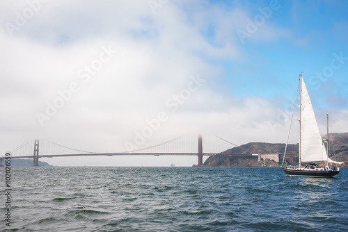 Tranquil scene of San Francisco Bay with white sailboat under Golden Gate Bridge in light mist. Reflective water and rolling hills in backdrop. Typical tourist spot.