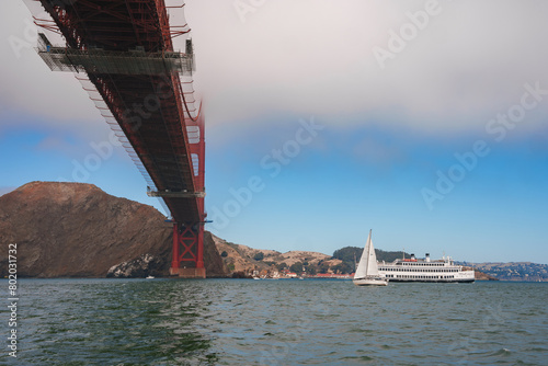 Iconic Golden Gate Bridge from below, towering red structure against blue sky in San Francisco Bay. Yacht sailing leisurely, Marin Headlands backdrop. photo