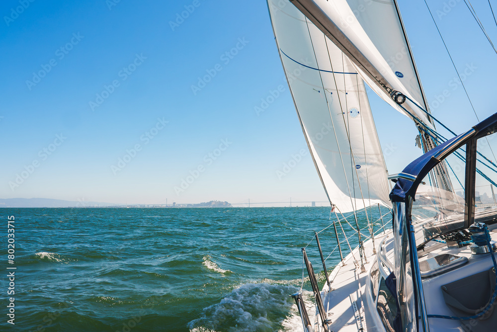 Fototapeta premium Sailboat sailing on serene waters near San Francisco, white sails billowing under a clear blue sky. Modern yacht deck and rigging visible, Bay Bridge in background.
