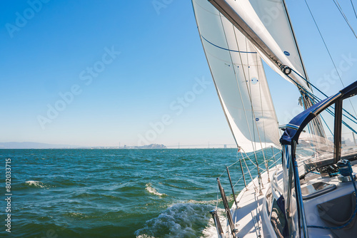 Sailboat sailing on serene waters near San Francisco, white sails billowing under a clear blue sky. Modern yacht deck and rigging visible, Bay Bridge in background. photo