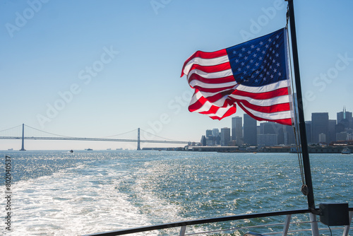 American flag fluttering on boat as it leaves SF towards Bay Bridge. Skyline, buildings, river wake visible on sunny day. Maritime culture essence captured.
