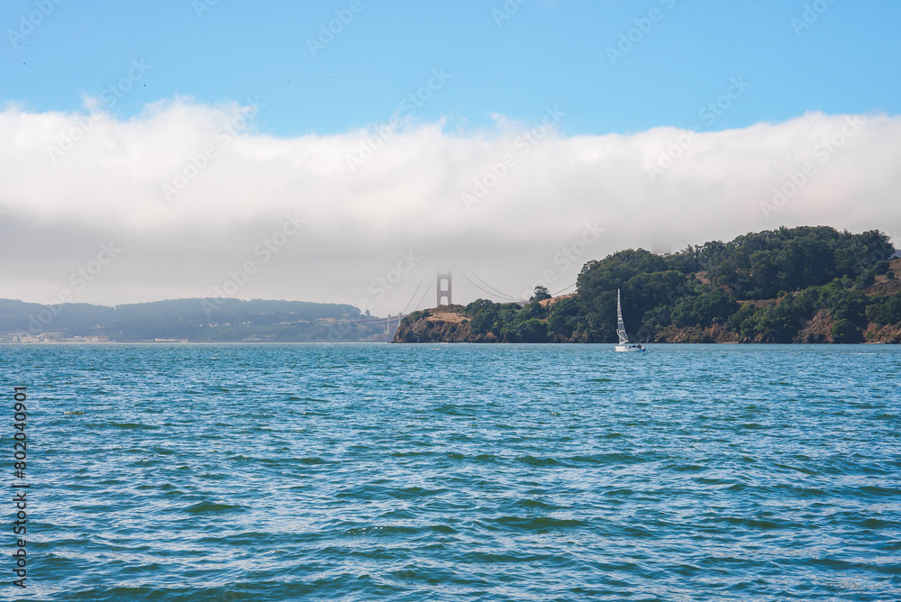 Serene view of San Francisco Bay with partially shrouded Golden Gate Bridge in mist. Blue waters, white sailboat, green landmass, and cloudy sky add to tranquil beauty. Ideal for travel blogs.