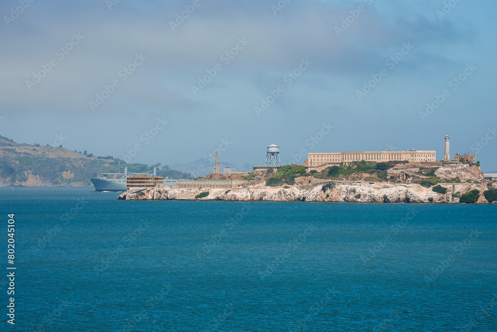 A captivating view of Alcatraz Island in San Francisco Bay. The infamous former prison stands out against the skyline, with a hazy sky and rolling hills in the background.