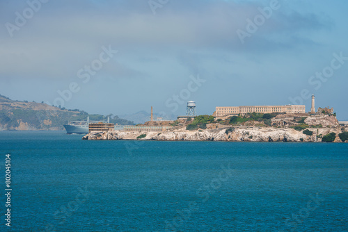 A captivating view of Alcatraz Island in San Francisco Bay. The infamous former prison stands out against the skyline, with a hazy sky and rolling hills in the background.