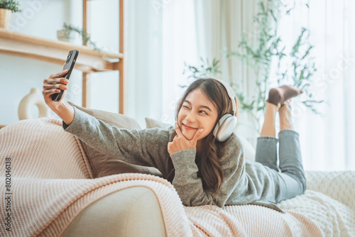 Smiling asian woman with headphones taking selfie with mobile smartphone on couch at home © oatawa