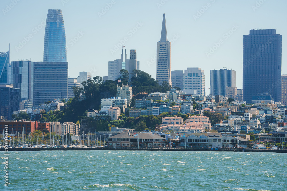 Fototapeta premium Scenic view of San Francisco's waterfront with glistening bay waters, yachts, historic pier buildings, and modern city skyline featuring iconic skyscrapers.