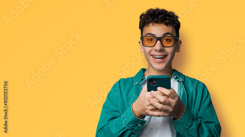 Excited surprised shocked astonished happy curly haired funny young man wear braces sunglasses glasses open mouth hold typing cell phone cellular smartphone, isolated yellow background photo