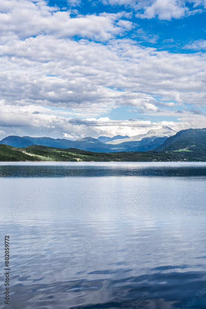 Overlooking the flat, calm waters of the Ålvundfjord. View of the other side of the fjord coastline with the village of Handlarstuhamran against the backdrop of the Trollheimen Mountains in Norway.