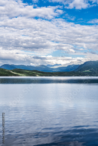 Overlooking the flat, calm waters of the Ålvundfjord. View of the other side of the fjord coastline with the village of Handlarstuhamran against the backdrop of the Trollheimen Mountains in Norway.