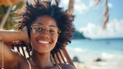 Portrait of happy young black woman relaxing on wooden deck chair at tropical beach while looking at camera wearing spectacles. Smiling girl with fashion sunglasses enjoying vacation