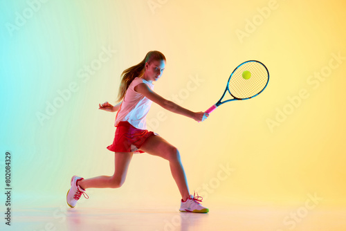 Portrait of young sportive teenager, female tennis player in uniform training in motion in neon light against blue-yellow background. Concept of individual kind of sports, fashion, tournament, action © Lustre