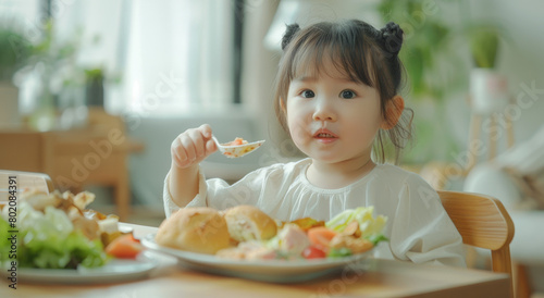 A cute little Asian girl is sitting at the dining table, eating her lunch with one hand and holding an open sandwich with the other hand.