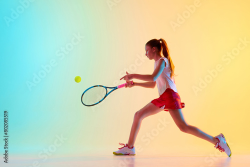 Fit teen girl, tennis athlete trains skills of hitting ball from opponent in neon light against blue-yellow background. Concept of individual kind of sports, fashion, tournament, action. Ad © Lustre