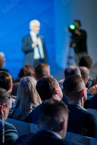 Focused view of a businessman delivering a speech at a corporate conference with an attentive audience and a videographer capturing the event.