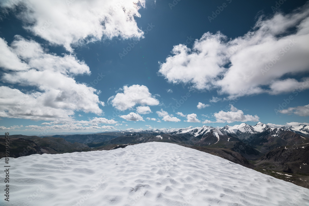 Obraz premium Epic vast top view from snow-capped vertex to wide alpine vastness. High mountains silhouettes with snowy tops. Snowfield at summit in sunlight. Large mountain range with snow-white peaks under clouds