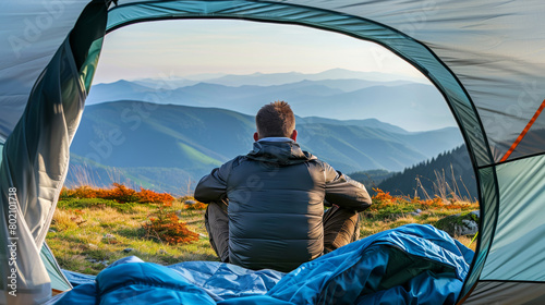 Back view of a man sitting in a tent, looking out at a panoramic mountain view during sunrise. photo