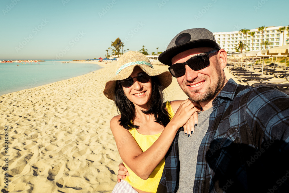 Happy multiracial couple taking selfie on summer vacation - Trendy man and woman having fun on Cyprus Nissi beach. Friendship and holidays concept with guy and girl enjoying summer