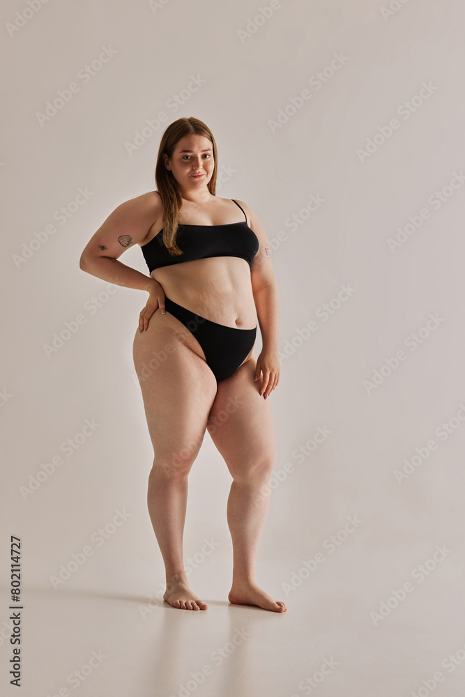 Full-length image of beautiful young girl with chubby body shape posing in black underwear against grey studio background. Concept of natural beauty, body positivity, care, acceptance