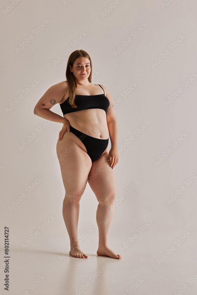 Full-length image of beautiful young girl with chubby body shape posing in black underwear against grey studio background. Concept of natural beauty, body positivity, care, acceptance Full-length image of beautiful young girl with chubby body shape posing in black underwear against grey studio background. Concept of natural beauty, body positivity, care, acceptance