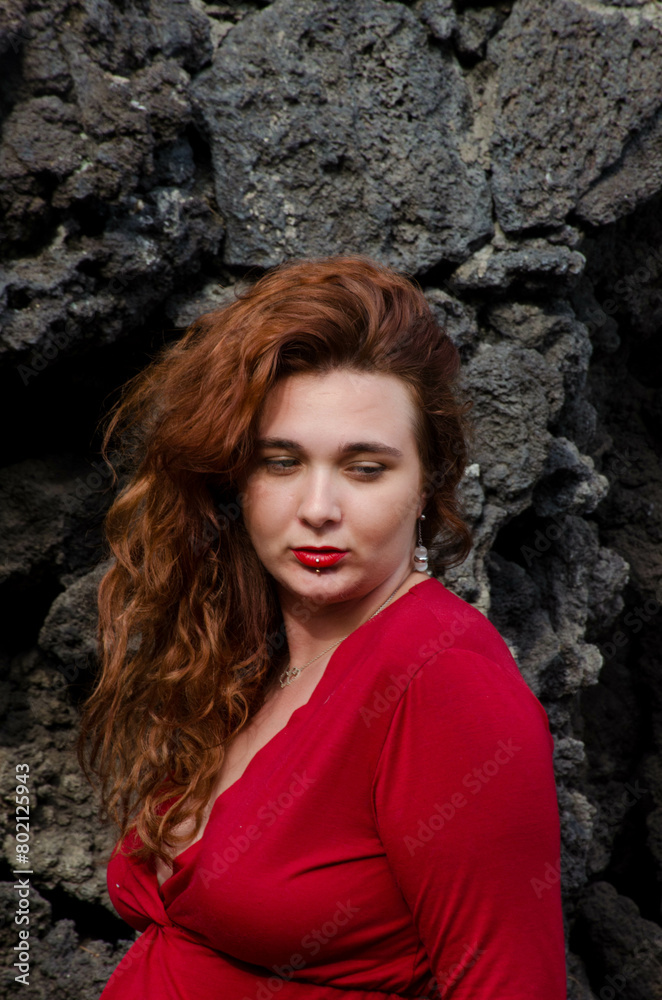 Portrait de femme rousse avec une robe rouge devant un mur en pierres noires Portrait de femme rousse avec une robe rouge devant un mur en pierres noires
